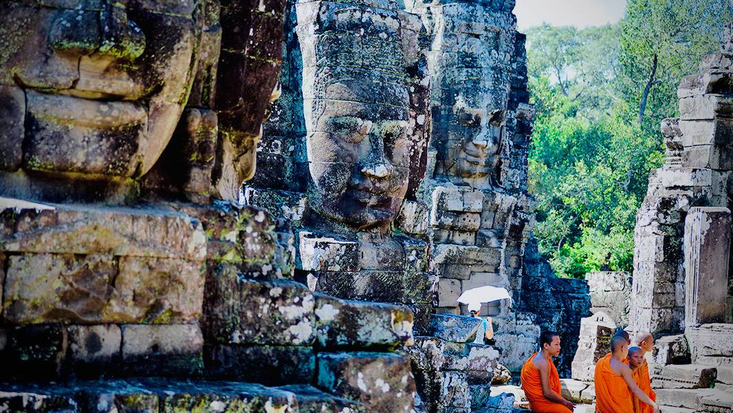 Buddhist monks in saffron robes sit beneath the iconic smiling stone faces of Bayon Temple, the spiritual heart of Angkor Thom in Siem Reap, Cambodia.